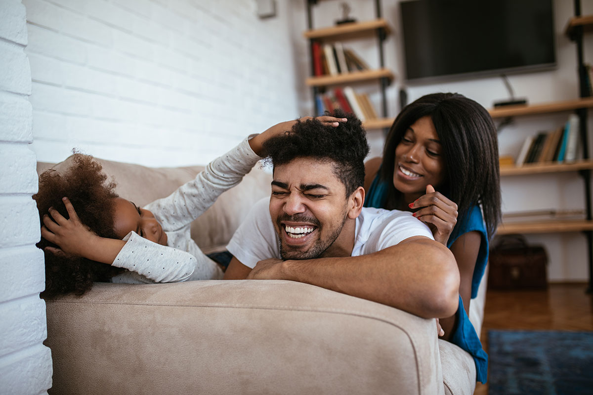 Two adults and a child playing on a sofa in a room with exposed brick walls painted white, and shelves containing books and a screen in the background